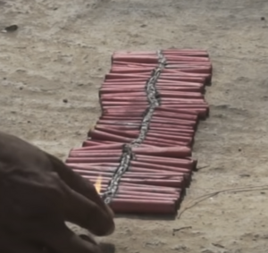 A screen capture of a video showing a hundred bijili crackers strung together being lit